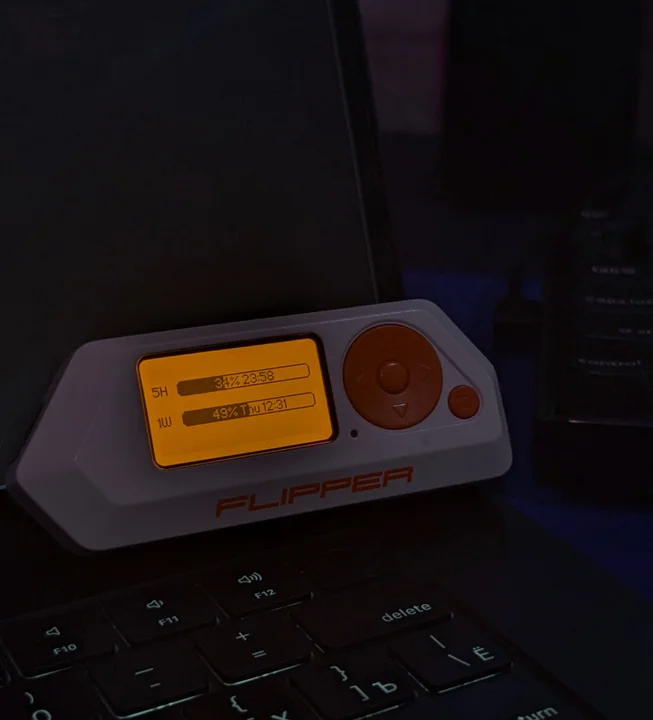 A Flipper Zero resting on a keyboard, with 5H and 1W usage bars on its orange screen.